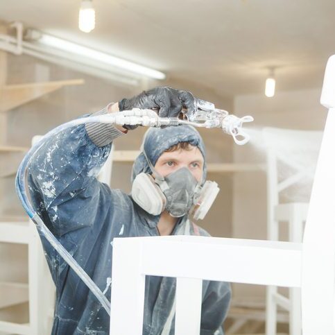 Man painting chair into white paint in respiratory mask. Application of flame retardant ensuring fire protection, airless spraying. Man painting chair into white paint in respiratory mask. Application of flame retardant ensuring fire protection, airless spraying device.