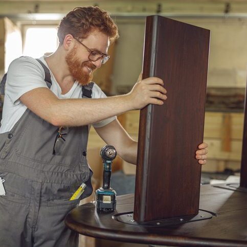 Smiling carpenter fixing table with electric drill standing in carpentry workshop Woodworker fixing table with electric drill standing in carpentry workshop. High quality photo